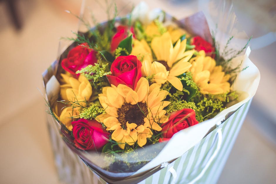 A vibrant floral arrangement featuring yellow sunflowers with dark centers, bright pink roses, and small green filler flowers, all arranged in a bouquet wrapped in clear cellophane and white tissue pa