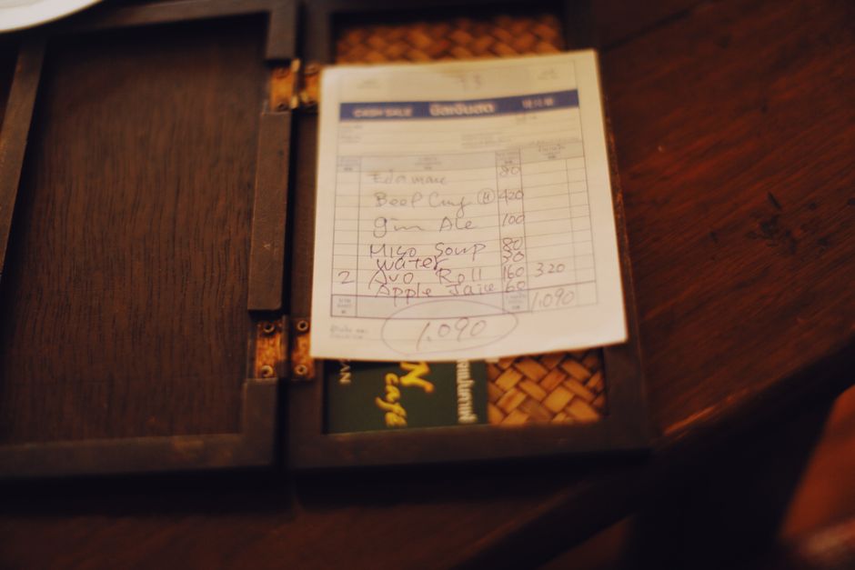 A handwritten restaurant bill placed inside a dark wooden menu cover on a wooden table, with visible metal hinges and a woven pattern underneath the menu. The bill lists food items such as calamari, b
