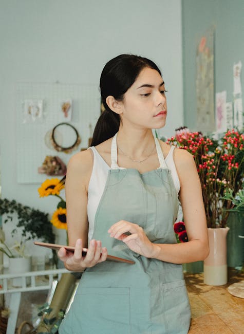 A young woman with long dark hair pulled back is standing inside a flower shop, holding a smartphone in her left hand. She is wearing a white sleeveless top and a light green apron, indicating she is 