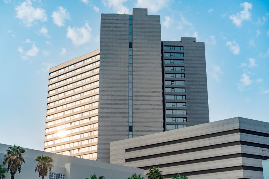 A modern high-rise office building with a sleek, light-colored exterior, featuring horizontal and vertical lines created by windows and structural panels. The building stands against a bright blue sky