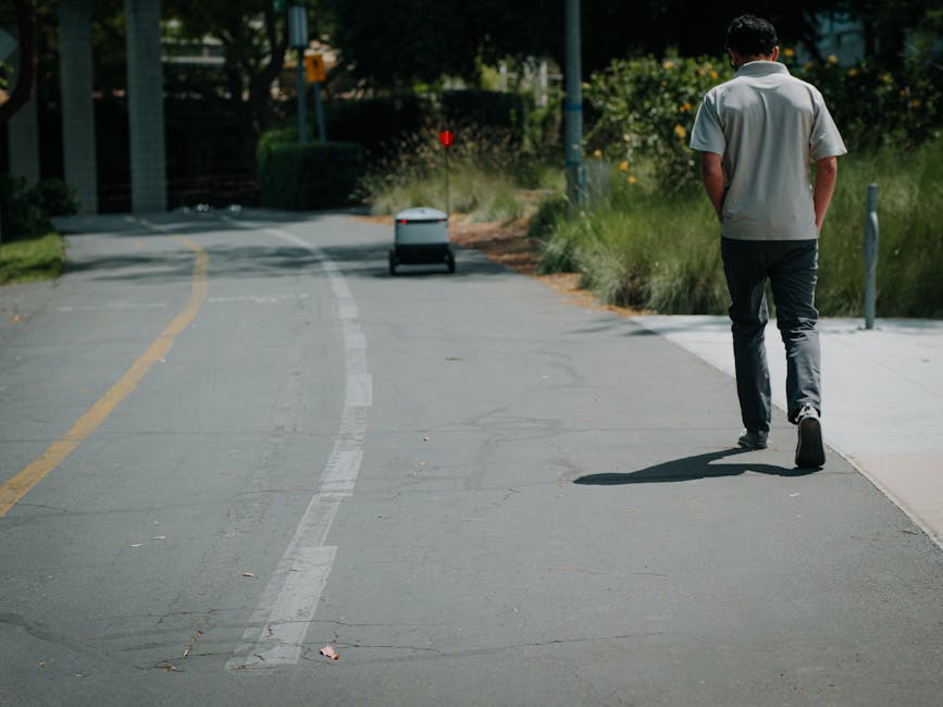 A man dressed in casual grey and black clothing is walking along a paved sidewalk next to a bicycle lane, with a delivery robot approaching in the distance. The scene is set in an urban area with gree