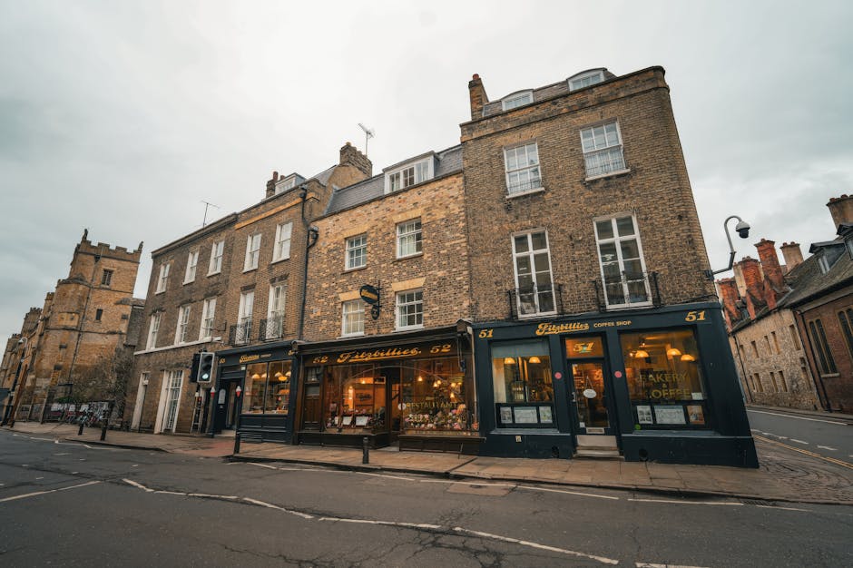 A charming street scene featuring a flower shop with a black façade and gold lettering, located in a historic brick building with multiple floors and white window frames. The shop's large glass windo