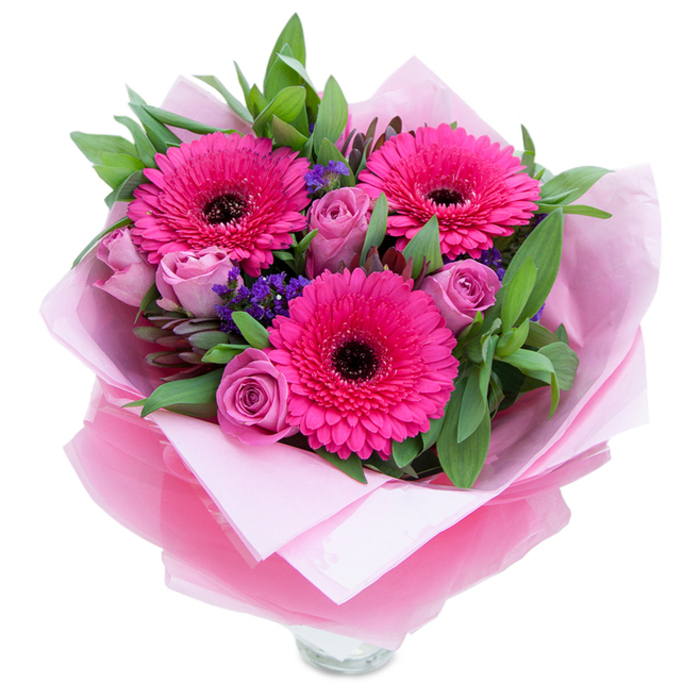 A graceful, pink-hued bouquet that balances playful energy with quiet elegance, pictured as if placed on a small café table by Bath Road in Sipson. Three radiant Gerbera daisies in an assertive fuchsia tone take centre stage, their layered petals forming a sunburst around rich dark centres that give depth and contrast. Soft lavender-pink roses nestle among them, petals tightly curled in the classic bud form that signals the blooms are newly cut and will open slowly, releasing gentle fragrance. Intermittent sprays of purple-blue statice add a textured, wildflower quality-small, clustered blossoms that lift the composition and introduce a complementary cool note. Lush green foliage, possibly salal or rounded eucalyptus leaves, frames and supports the flowers, the leaves broad and glossy with a slightly dewy sheen that suggests freshness straight from the cooler. The bouquet is wrapped in a matte pale-pink tissue inner layer and an outer sheet of translucent fuchsia-pink cellophane with a faint crinkle; the layers are folded with care so the arrangement reads as both celebratory and refined. At the lower edge, the top rim of a clear glass vase peeks through, indicating the bouquet is arranged for immediate display-ideal for brightening a hall table or centrepiece at a Sipson gathering. The scent is light and inviting, roses offering a soft perfume and gerberas a subtle green sweetness. This piece feels perfect for birthdays, Mother's Day, or an anniversary where warmth and thoughtfulness are the message.